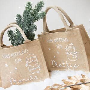 May include: Tan burlap tote bags with white text and illustrations. The bags have handles and feature holiday-themed designs, including a snowman, Santa Claus, and a bunny. The text on the bags reads "Vom Nikolaus F&uuml;r" and includes names.