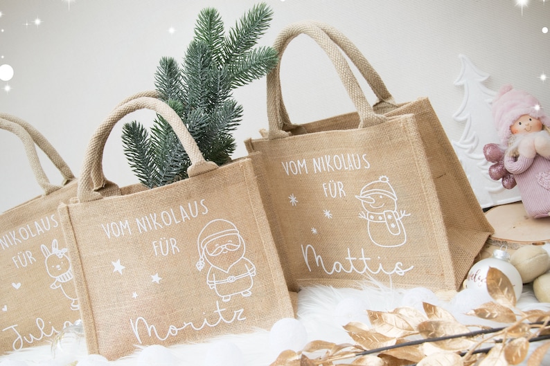May include: Tan burlap tote bags with white text and illustrations. The bags have handles and feature holiday-themed designs, including a snowman, Santa Claus, and a bunny. The text on the bags reads "Vom Nikolaus F&uuml;r" and includes names.