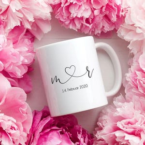 Op de afbeelding: Een witte keramische mok met een handvat, met een hart en de letters "m r" in zwart schrift, samen met de datum "14. Februar 2020". De mok is omgeven door roze pioenrozen.