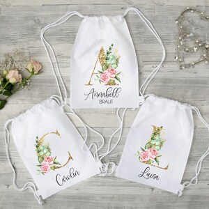 Może przedstawiać: Trzy białe worki sznurkowe z literami z błyszczącego złota i wzorami kwiatowymi. Worki są spersonalizowane z nazwami Annabell Braut, Carolin i Luisa.