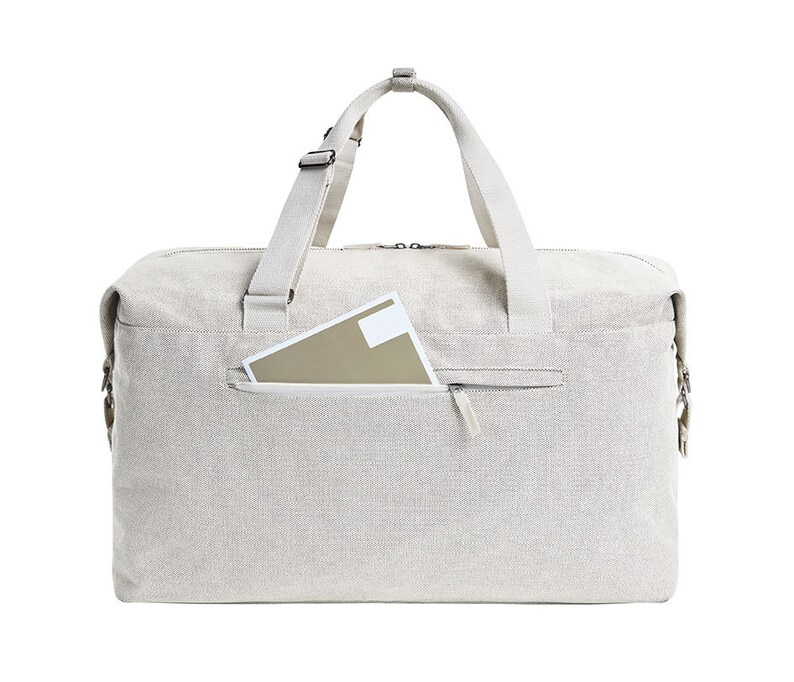 Puede incluir: Una bolsa de lona beige con un bolsillo con cremallera en la parte delantera. La bolsa tiene un solo compartimento con cremallera y dos asas superiores.