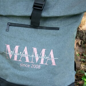 May include: Gray canvas backpack with a black zipper and buckle. The backpack has a pink glitter design that says "MAMA" with the names "Martina" and "Nicola" and the text "since 2008".