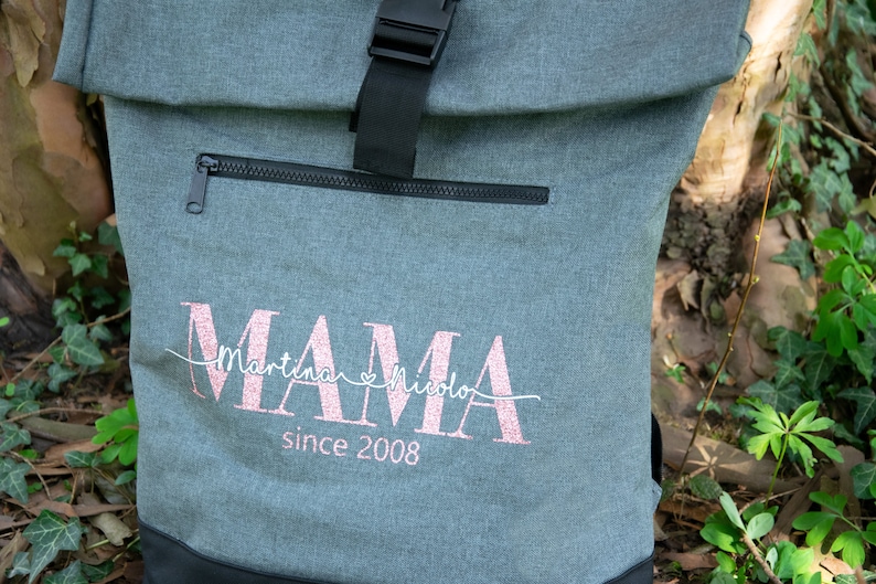 May include: Gray canvas backpack with a black zipper and buckle. The backpack has a pink glitter design that says "MAMA" with the names "Martina" and "Nicola" and the text "since 2008".