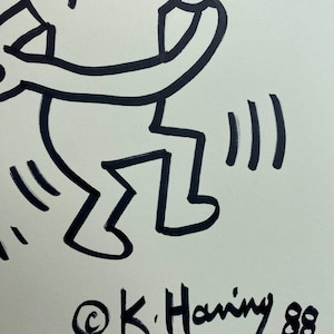 May include: Black and white line drawing of a dancing figure with raised arms. The artwork is signed "&copy;K. Haring 88" in black ink. The figure has a simple, bold outline, and the style is reminiscent of street art.