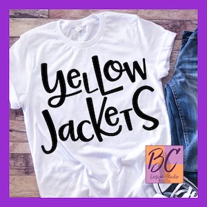 Könnte beinhalten: Weißes T-Shirt mit schwarzem Text, der "Yellow Jackets" lautet. Das Shirt liegt auf einer Holzoberfläche mit einer blauen Jeans und einem weißen Schuh im Hintergrund. Das Bild enthält auch ein Logo unten rechts, das "BC Design Studio" lautet.