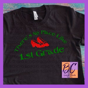 May include: A black t-shirt with green text that reads "There's No Place Like 1st Grade". Red glitter ruby slippers are above the text. The text "BC Design Studio" is in the lower right corner.