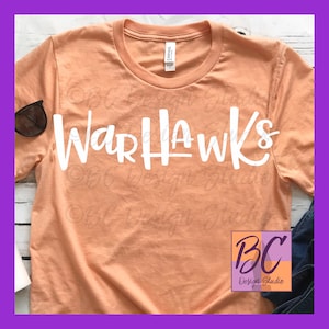 May include: A peach-colored t-shirt with the text "Warhawks" in white lettering. The shirt is laid flat on a white surface with a pair of sunglasses and a pair of blue jeans in the background.