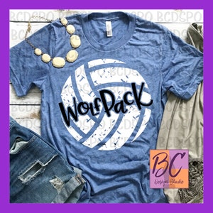 Pode incluir: Uma camiseta azul com um gráfico de voleibol branco e o texto "Wolf Pack" em preto. A camiseta está sobre uma superfície de madeira com um par de calças de ganga e uma camisa castanha.