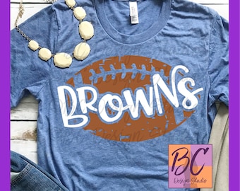 Könnte beinhalten: Ein blaues T-Shirt mit einem braunen Football-Motiv und dem Wort "Browns" in weißer Schrift. Das Football-Motiv hat weiße Nähte und einen Used-Look.
