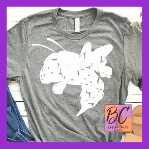 May include: A gray t-shirt with a white distressed graphic of a bee. The graphic is in the center of the shirt. The shirt is laying on a white surface. The text "BC Design Studio" is in the lower right corner of the image.