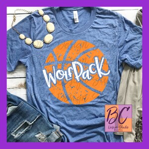 Pode incluir: Uma camiseta azul com um gráfico de basquete laranja e o texto "Wolf Pack" em branco. A bola de basquete tem um visual desgastado.