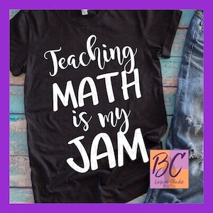 May include: Black t-shirt with white text that reads "Teaching Math is my Jam". The shirt is on a wooden surface with a pair of blue jeans and a logo that reads "BC Design Studio".