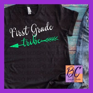 Pode incluir: Camiseta preta com texto branco que diz "First Grade Tribe" e uma flecha verde com penas apontando para a direita. A camiseta está sobre uma superfície de madeira azul.