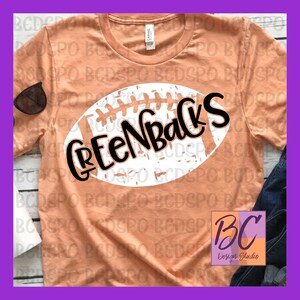 Puede incluir: Una camiseta de color melocotón con un gráfico de fútbol blanco desgastado que dice "Greenbacks" en letras negras.