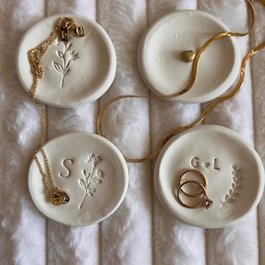 May include: Four small, round, white ceramic trinket dishes with gold jewellery. Two dishes have floral designs, one has a heart, and one has initials. Gold necklaces and rings are displayed on the dishes.
