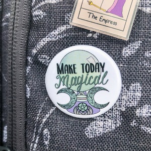 May include: A gray canvas bag with a white floral pattern and a zipper. A round button with the text "Make Today Magical" is attached to the bag. The button has a green, purple, and white design with a crescent moon and a pentagram.