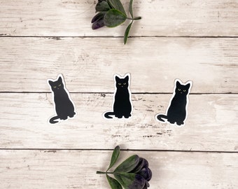 Puede incluir: Tres pegatinas de gatos negros sobre un fondo de madera clara. Los gatos están sentados con las colas enroscadas detrás de ellos. Hay dos pequeñas plantas verdes en el fondo.
