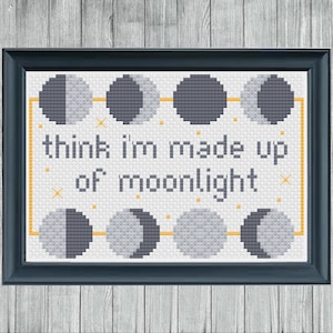 May include: A framed cross-stitch artwork with a dark blue frame. The design features phases of the moon and the text "think I'm made up of moonlight" in a simple font. The background is white with a yellow border.