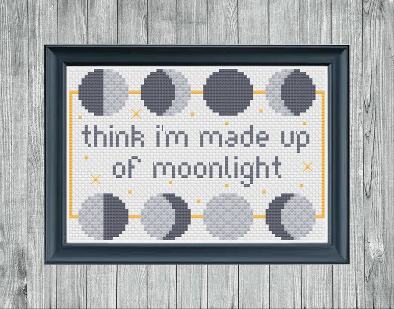 May include: A framed cross-stitch artwork with a dark blue frame. The design features phases of the moon and the text "think I'm made up of moonlight" in a simple font. The background is white with a yellow border.
