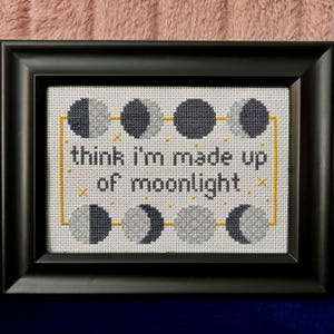 May include: Cross-stitch art in a black frame. The design features phases of the moon in gray and navy blue, connected by yellow lines and accents. The text "think I'm made up of moonlight" is stitched in black.