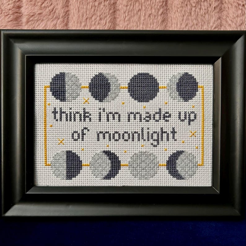 May include: Cross-stitch art in a black frame. The design features phases of the moon in gray and navy blue, connected by yellow lines and accents. The text "think I'm made up of moonlight" is stitched in black.