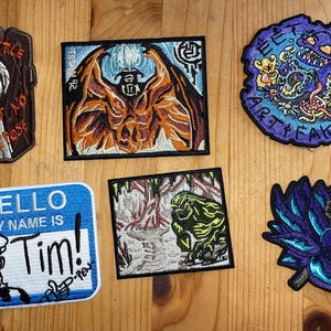 Peut inclure: Collection de patchs brodés avec divers motifs. Comprend un patch en forme de cercueil avec du texte, une créature ailée, un patch avec le texte "HELLO MY NAME IS Tim!", un monstre, une scène de forêt et une fleur violette.