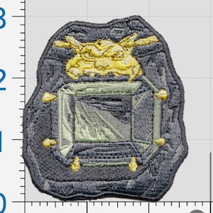 May include: An embroidered patch featuring a grey stone-like border with a yellow creature head at the top and a central rectangular opening. The patch is positioned against a ruler showing a height of approximately 2.5 inches.