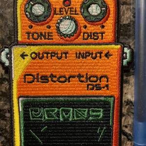 Pode incluir: Patch bordado com um pedal de efeitos de guitarra laranja e amarelo. O patch inclui os textos "CHECK", "LEVEL", "TONE", "DIST", "OUTPUT", "INPUT" e "Distortion DS-1". Apresenta detalhes em verde e preto.