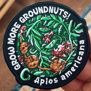 May include: A black and green embroidered patch with the text "Grow More Groundnuts! Apios Americana" in white. The patch features a green vine with brown peanuts and pink flowers.