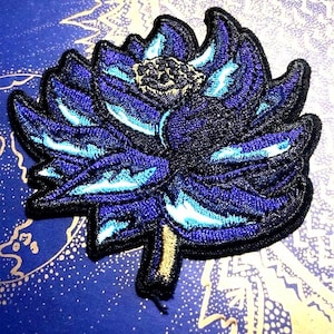 Puede incluir: Un parche bordado de una flor azul y blanca con un tallo amarillo. La flor tiene un diseño detallado con múltiples capas de pétalos.