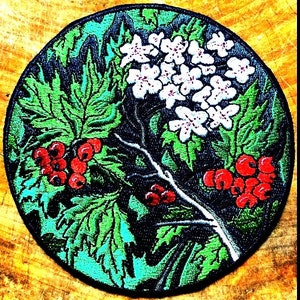 May include: A round embroidered patch with a green, white, and red floral design. The patch features a branch with white flowers and red berries, surrounded by green leaves. The background is black.