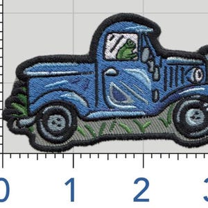 May include: Embroidered patch featuring a blue vintage pickup truck with a frog driver. The truck has black detailing and sits on a bed of green grass. The patch is approximately 3 inches long.