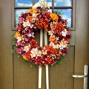 Peut inclure: Une couronne florale circulaire composée d'un arrangement dense de fleurs artificielles dans des tons de bordeaux profond, orange brûlé, jaune doré et crème. La couronne comprend de petits accents de feuillage vert et deux longs rubans beige clair.