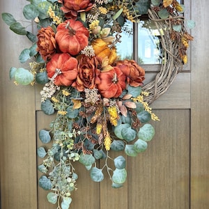 May include: A decorative wreath featuring orange velvet pumpkins, deep rust-coloured flowers, and green eucalyptus leaves arranged on a natural twig base. The arrangement includes small yellow floral accents and brown pinecone-like elements.