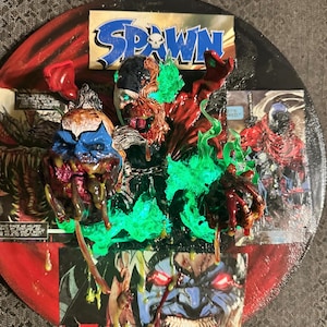 Könnte beinhalten: Ein rundes, mehrschichtiges Kunstwerk mit der Comicfigur Spawn. Die zentrale Figur ist von grünen Flammen und Comic-Panels umgeben. Das Wort "SPAWN" ist oben zu sehen. Das Kunstwerk hat ein rot-schwarzes Farbschema.