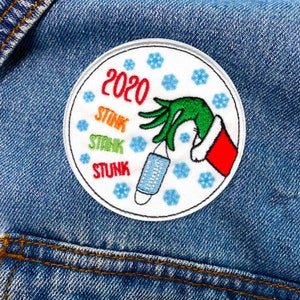 Op de afbeelding: Een ronde, witte geborduurde patch met een groene hand die een blauw en wit mondkapje vasthoudt. De hand draagt een rode kerstmanmuts. De tekst "2020" staat bovenaan de patch, en het woord "STUNK" wordt drie keer in verschillende kleuren onder de hand herhaald.
