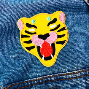 Könnte beinhalten: Ein gestickter gelber Tigerkopf-Patch mit schwarzen Streifen, rosa Nase und Mund und roter Zunge. Der Tiger hat zwei blaue Sterne auf der Stirn.
