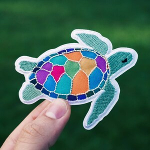 May include: A colourful embroidered patch of a sea turtle with a stained glass design. The turtle is outlined in white and has a green shell.
