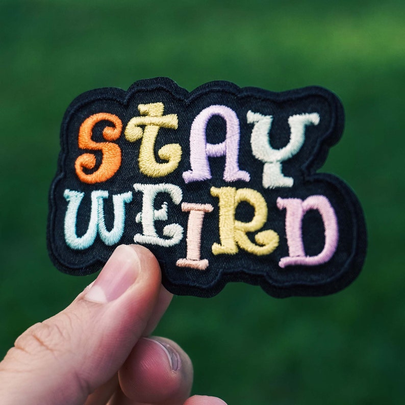 Puede incluir: Parche bordado con las palabras "STAY WEIRD" en letras coloridas. El parche tiene un borde negro y se sostiene sobre un fondo verde borroso. Las letras son de color naranja, amarillo, rosa, azul y blanco.