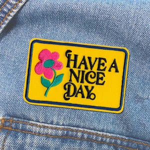 May include: A yellow embroidered patch with a pink flower and the text "Have a Nice Day" in black lettering.