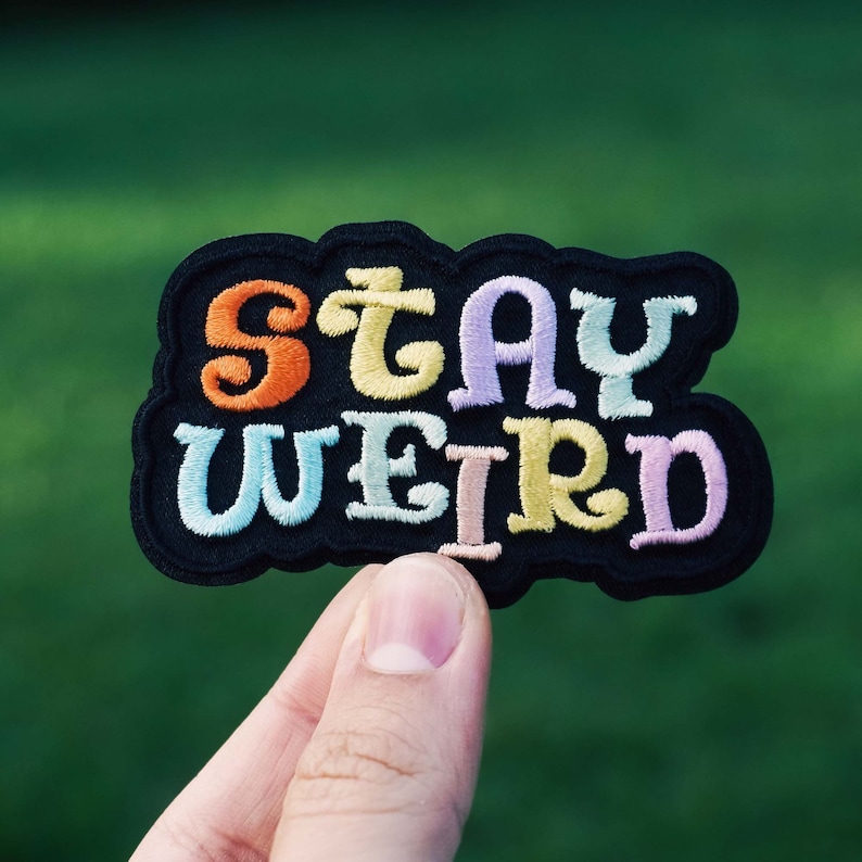 Puede incluir: Un parche bordado negro con las palabras "STAY WEIRD" en letras coloridas estilo burbuja. Las letras son de color naranja, amarillo, azul claro, morado y rosa. El parche tiene un borde negro y se sostiene sobre un fondo verde borroso.