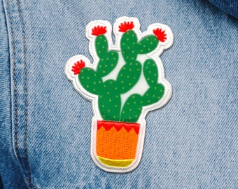 Puede incluir: Un cactus verde con flores rojas en una maceta amarilla y naranja. El cactus está bordado en un parche de tela blanco.
