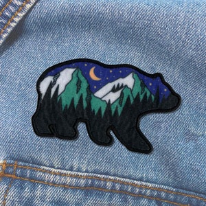May include: A black bear silhouette with a mountain range and a crescent moon inside. The mountains are green and white with a starry night sky background.
