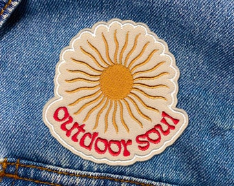 Może przedstawiać: Haftowana naszywka z motywem słońca w kolorze żółtym i pomarańczowym, z napisem "Outdoor soul" w kolorze czerwonym. Naszywka znajduje się na niebieskiej kurtce dżinsowej.