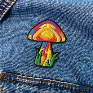 May include: A colorful embroidered patch of a mushroom with a yellow cap, red and orange accents, and a green stem. The patch is on a blue denim background.