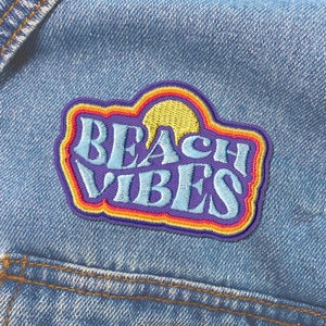 May include: A blue denim jacket with a colorful embroidered patch that says "Beach Vibes". The patch has a yellow sun with orange, red, and purple outlines.