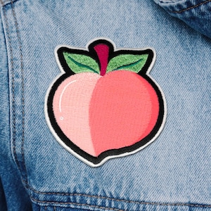 Könnte beinhalten: Ein gestickter Pfirsich-Patch in Rosa und Weiß mit grünen Blättern. Der Patch befindet sich auf einem blauen Jeans-Hintergrund.