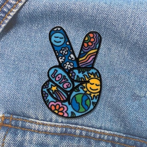 May include: A fabric patch shaped like a peace sign hand gesture, featuring a blue background with colorful illustrations including a smiling sun, a globe, flowers, a shooting star, and the word LOVE in pink letters, all outlined in black thread.