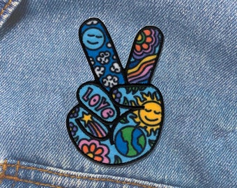 Könnte beinhalten: Ein Stoffaufnäher in Form eines Peace-Zeichens mit blauem Hintergrund und bunten Illustrationen, darunter eine lächelnde Sonne, ein Globus, Blumen, eine Sternschnuppe und das Wort LOVE in rosa Buchstaben, alles mit schwarzem Garn umrandet.