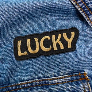 Op de afbeelding: Een zwarte stoffen patch met het woord "LUCKY" geborduurd in gouden draad. De patch is genaaid op een blauw denim jack.